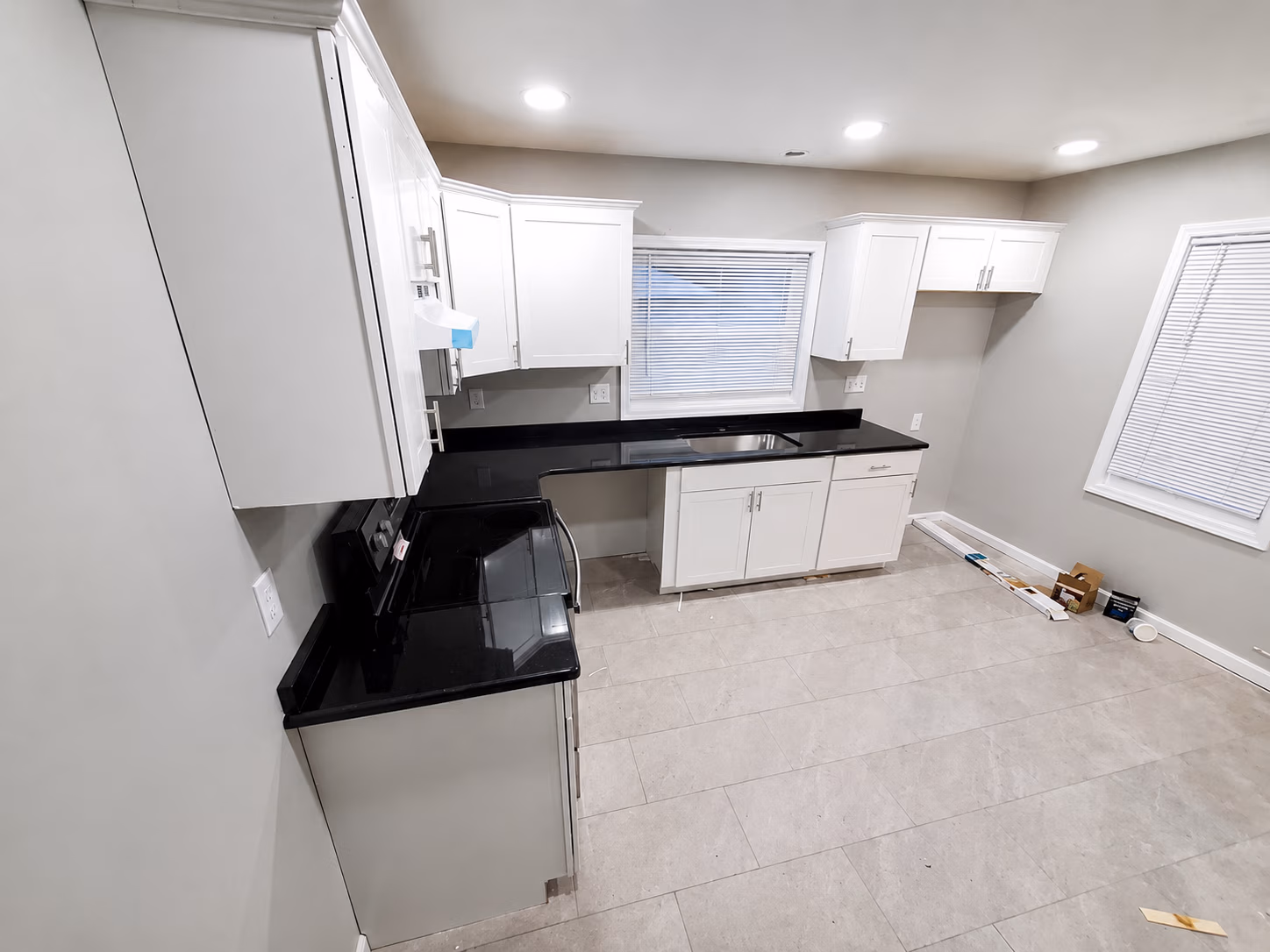 Modern empty kitchen with white cabinets, black countertops, tile floor, and two windows with blinds.